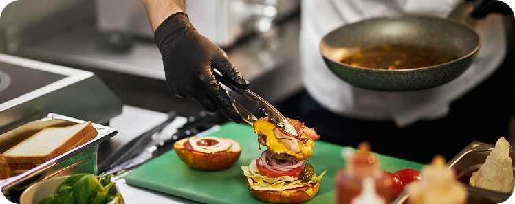 Food service worker assembling a burger with fresh ingredients at a commercial kitchen preparation station.