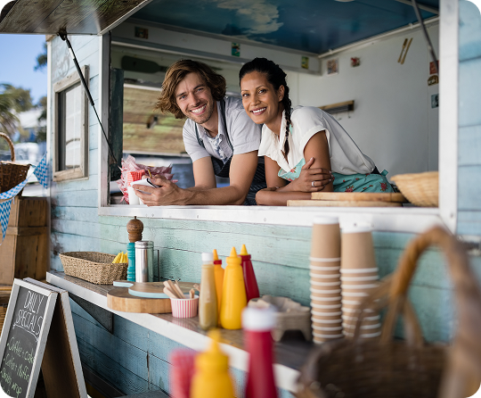 Food truck staff leaning from a service window above a counter with condiment bottles, disposable cups, and food service supplies.