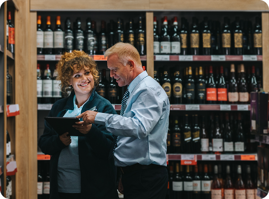 Store staff reviewing inventory data on a tablet in a liquor store aisle with wine and beverage shelves in the background.