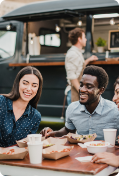 Guests enjoying meals and beverages at an outdoor dining table near a food service venue.